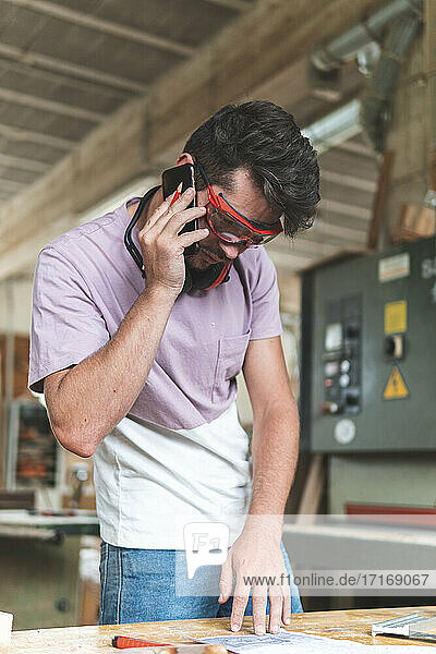 Young male craftsperson talking on mobile phone while working in workshop