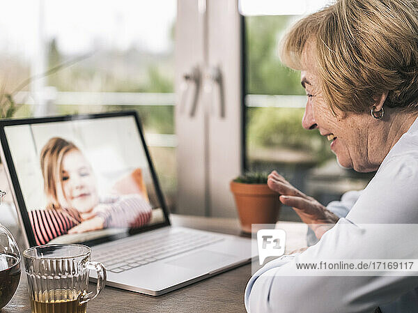 Lächelnde Großmutter  die ihrem Enkelkind bei einem Videogespräch über einen Laptop im Wohnzimmer zuwinkt