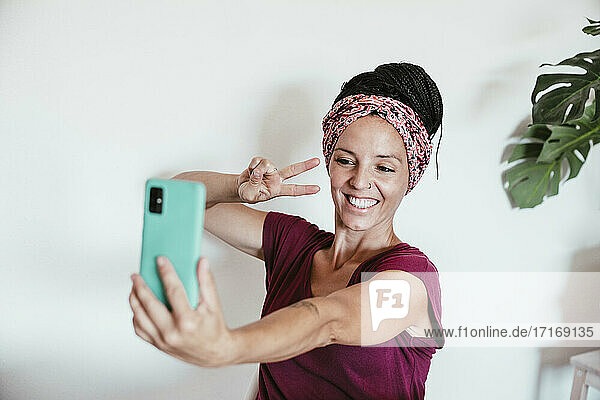 Lächelnde Frau  die ein Selfie mit ihrem Smartphone an der Wand zu Hause macht
