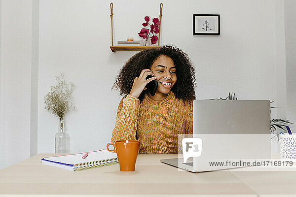 Lächelnde Frau mit Laptop  die im Büro zu Hause sitzt und mit ihrem Mobiltelefon spricht