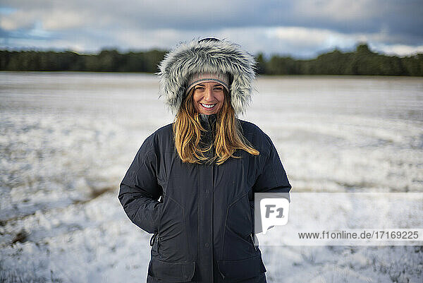 Lächelnde schöne Frau mit Händen in den Taschen  die auf einem verschneiten Feld vor dem Himmel steht