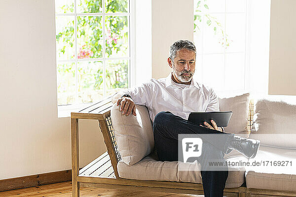Mature businessman using digital tablet while sitting with legs crossed at knee on sofa at home