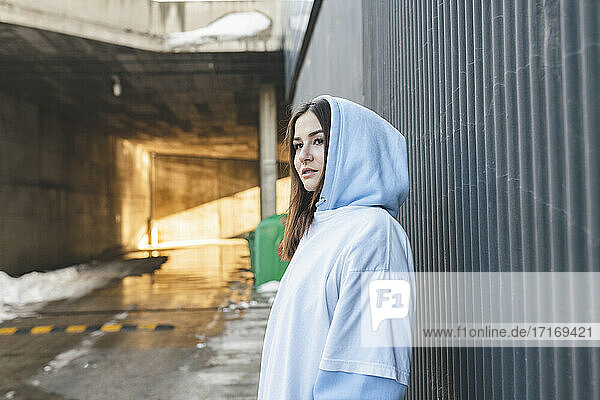 Young woman wearing hood clothing looking away while standing by wall