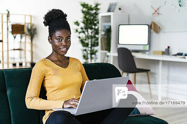 Smiling young Afro woman using laptop while sitting cross-legged on sofa in living room at home