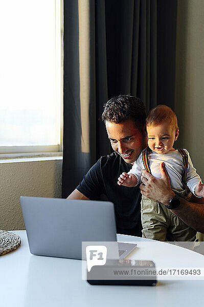 Mid adult man holding baby boy while working on laptop at home