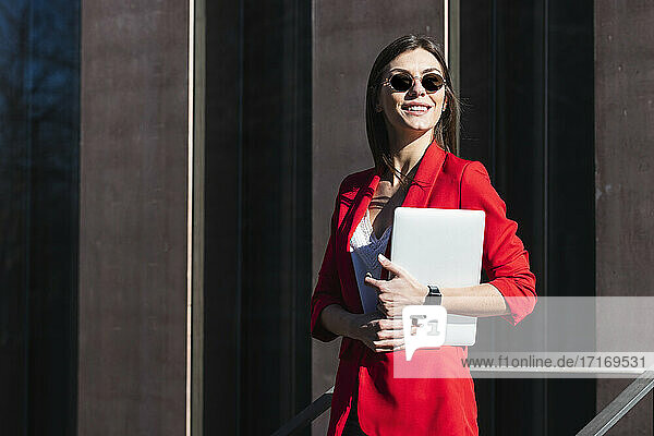 Smiling businesswoman with sunglasses holding laptop while standing against brown structure