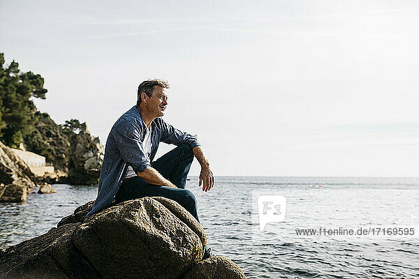 Thoughtful mature man sitting on rock against clear sky