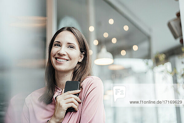 Smiling female freelancer holding smart phone while looking away in cafe