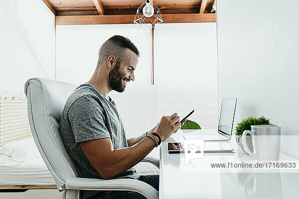 Lächelnder junger Mann  der zu Hause an Laptop und Mobiltelefon arbeitet