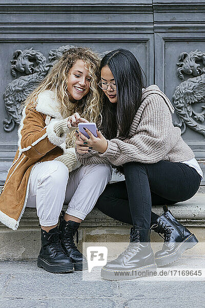 Young female friends using smart phone while sitting against door