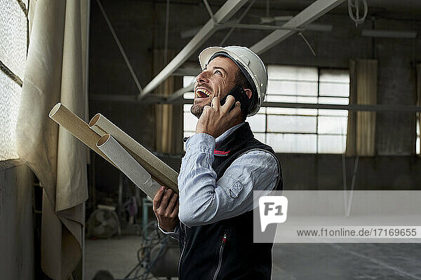 Cheerful male architect laughing while talking over mobile phone in constructing building