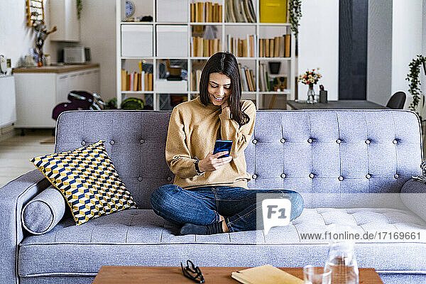 Young smiling woman using mobile phone while sitting on sofa at home