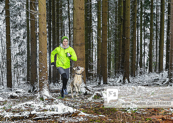 Junger männlicher Sportler joggt mit Labrador im Winter im Wald