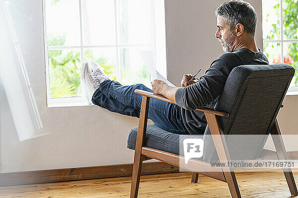 Geschäftsmann schreibt in Papier  während er mit gekreuzten Beinen auf einem Sessel zu Hause sitzt