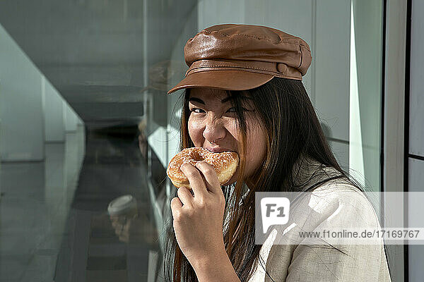 Young woman eating doughnut against glass wall