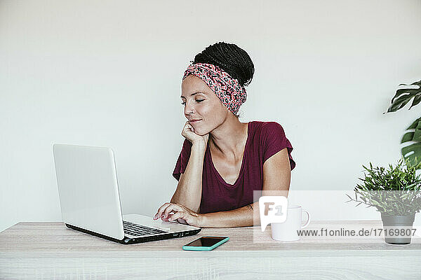 Weibliche Freiberuflerin arbeitet über einem Laptop mit der Hand auf dem Kinn gegen die Wand im Heimbüro