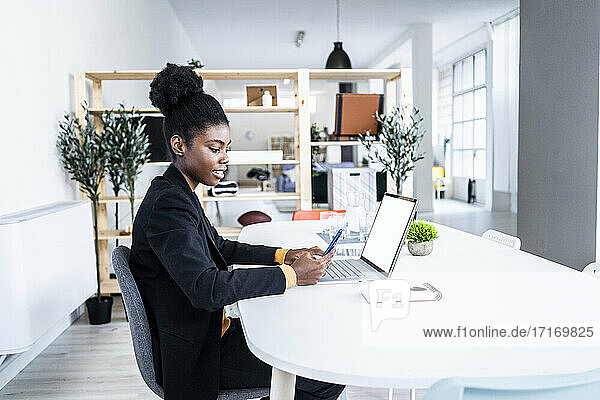 Young female Afro professional using smart phone while sitting with laptop at desk in office