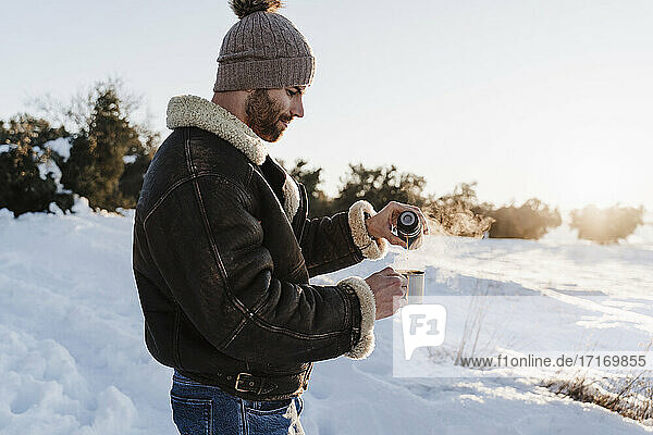 Young man with jacket pouring coffee in cup through thermos while standing on snow