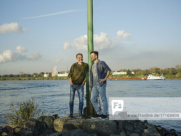 Smiling son and father leaning by pole while standing on rock against river