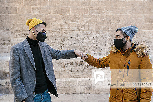 Junge Freunde mit Schutzmaske  die sich die Hände reichen  während sie an der Wand stehen