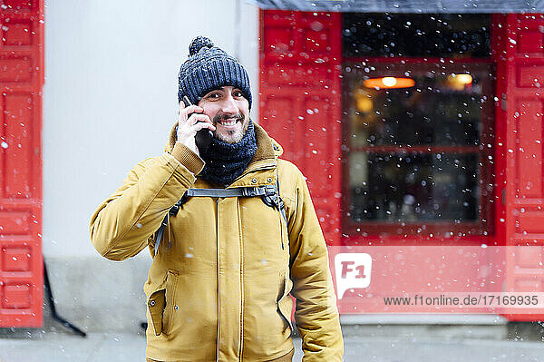 Smiling man talking on mobile phone while standing against store during winter