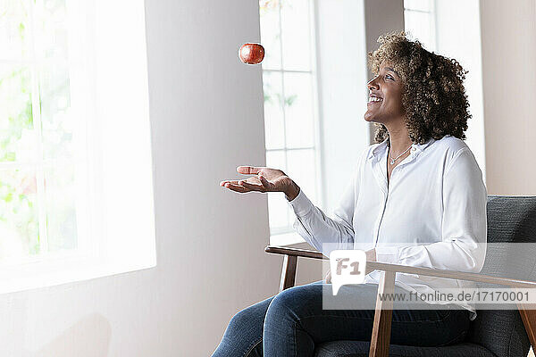 Smiling woman playing with apple while sitting on armchair at home