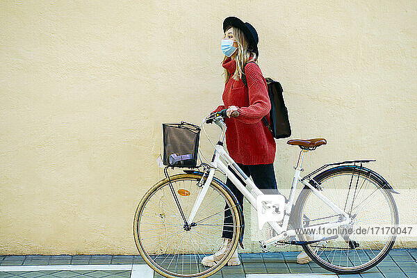 Young woman wearing face mask looking away while walking with bicycle on footpath