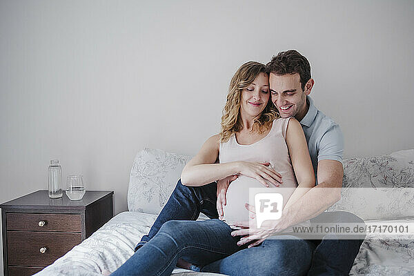 Lächelnder Mann berührt den Bauch seiner schwangeren Frau  während er zu Hause auf dem Bett sitzt