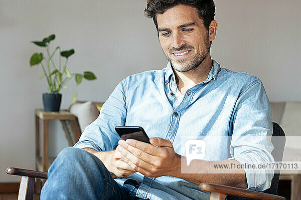 Mittelgroßer erwachsener Mann  der ein Mobiltelefon benutzt  während er zu Hause auf einem Sessel sitzt