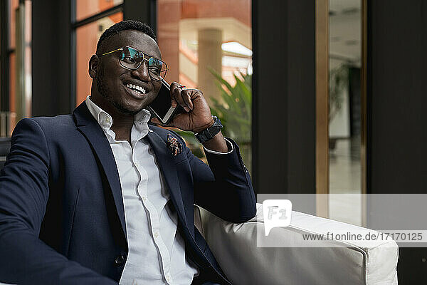 Lächelnder junger Geschäftsmann  der in einem Hotel sitzt und mit seinem Smartphone spricht