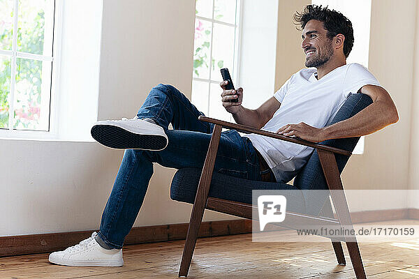 Lächelnder Mann  der ein Smartphone benutzt  während er sich zu Hause auf einem Sessel ausruht