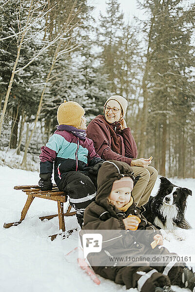 Lächelnde Mutter mit sitzender Tochter bei einem Wochenende mit Familie und Border Collie im Winter im Schnee