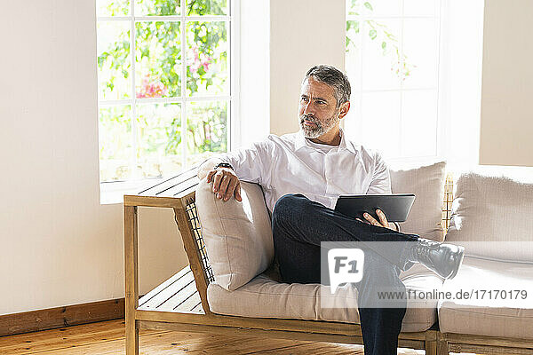Thoughtful businessman with digital tablet looking away while sitting on sofa at home