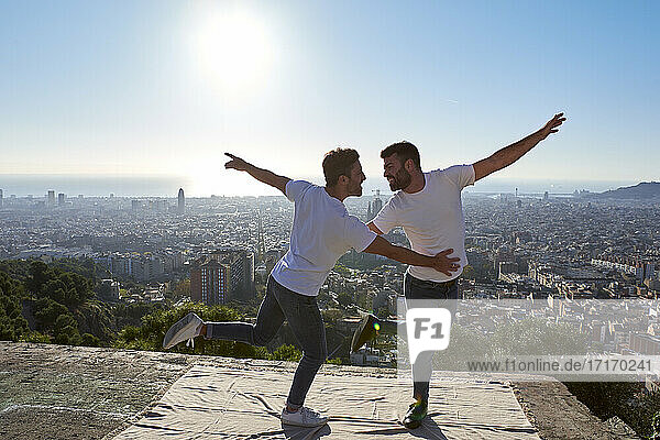 Happy gay couple with arms outstretched dancing on observation point against clear sky  Bunkers del Carmel  Barcelona  Spain