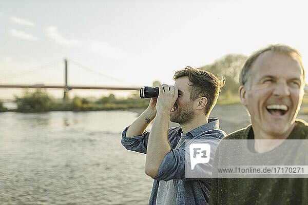 Son looking through binoculars while father laughing by at riverbank
