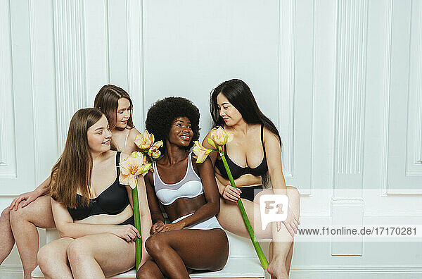 Lächelnde multiethnische Gruppe von Modellen in Dessous mit Blumen  die sich gegenseitig vor einer weißen Wand anschauen