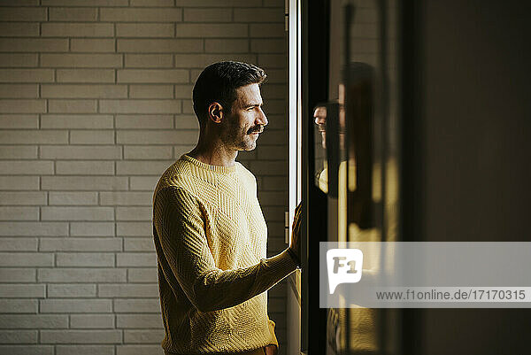 Man in yellow sweater looking through window at home