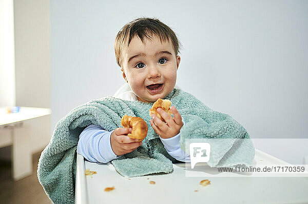 Cute baby wearing towel eating croissant while sitting by table at home
