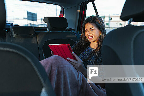 Lächelnde junge Frau  die ein digitales Tablet benutzt  während sie mit einer Decke auf dem Rücksitz eines Autos sitzt