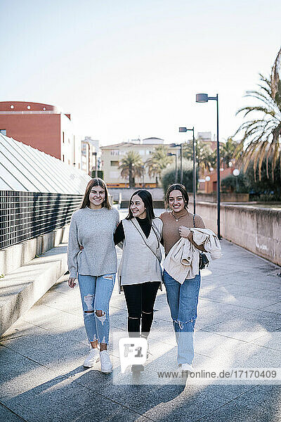 Smiling female friends walking on footpath against clear sky in city