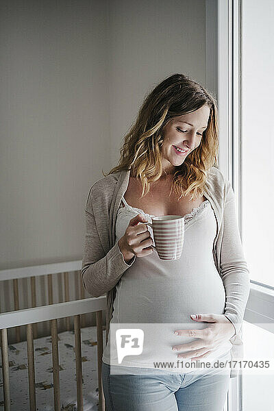 Lächelnde schwangere Frau mit Hand auf dem Bauch  die einen Kaffee hält  während sie zu Hause steht