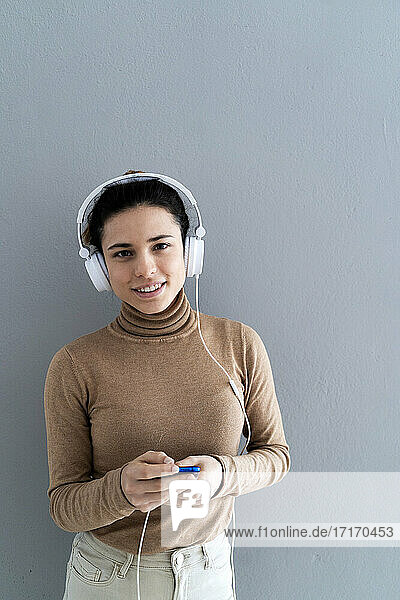 Lächelnde junge Frau  die ihr Smartphone hält  während sie Musik vor grauem Hintergrund hört