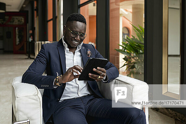 Smiling male entrepreneur working on digital tablet while sitting on armchair in hotel