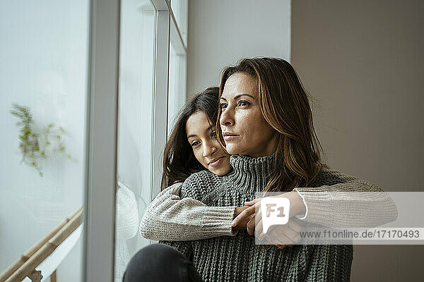 Tochter  die ihre Mutter umarmt  während sie zu Hause am Fenster nachdenkt