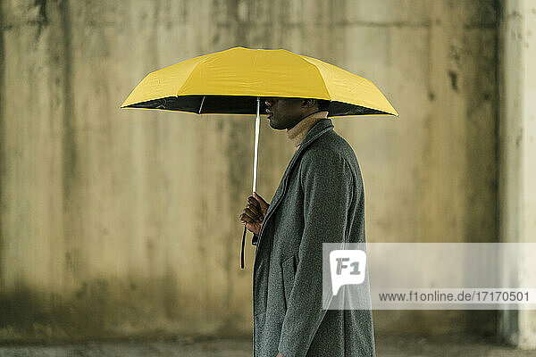 Junger Mann hält gelben Regenschirm  während er an der Wand steht
