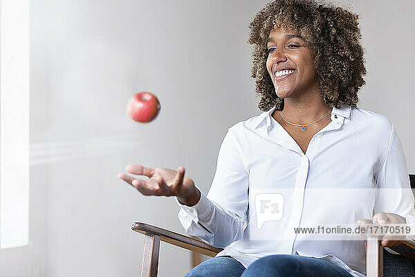 Frau lächelt  während sie mit einem Apfel spielt  der auf einem Sessel zu Hause sitzt