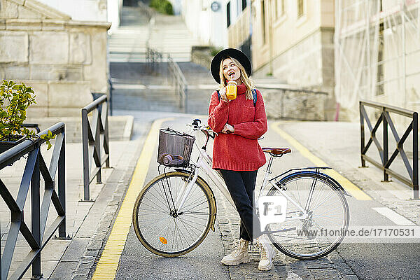 Young woman wearing hat drinking juice while standing with bicycle on road in city