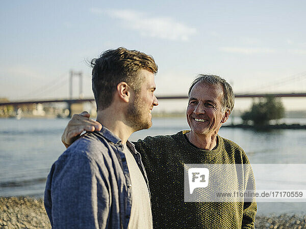 Happy father with arm around son at riverbank on sunny day