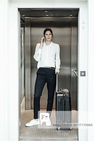 Unternehmerin mit Koffer  die im Aufzug stehend ihr Smartphone benutzt
