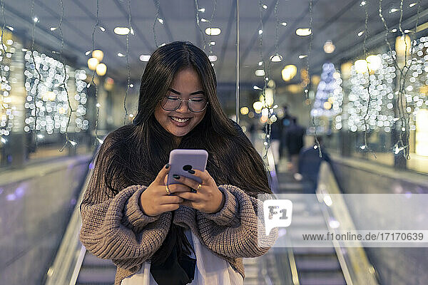 Young woman using smart phone while standing against illuminated glass wall in shopping mall